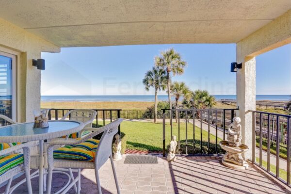 Covered patio with white wicker furniture, striped cushions, and garden statues overlooks a grassy yard, palm trees, and an ocean view under a clear blue sky.