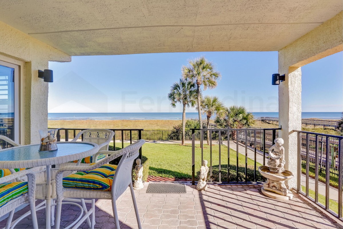 Covered patio with white wicker furniture, striped cushions, and garden statues overlooks a grassy yard, palm trees, and an ocean view under a clear blue sky.