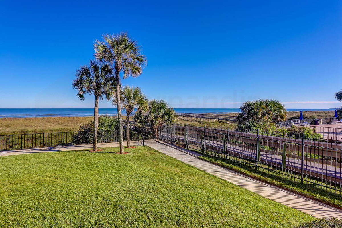 A grassy yard with palm trees, a black metal fence, and a boardwalk leading toward the beach and ocean under a clear blue sky.