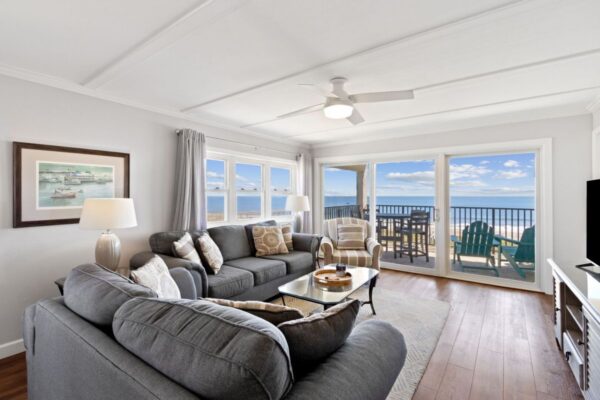 Bright living room with gray sofas, large windows, and glass doors opening to a balcony with ocean views. Cozy decor, lamp, wall art, and wooden floors create a relaxing beachside atmosphere.
