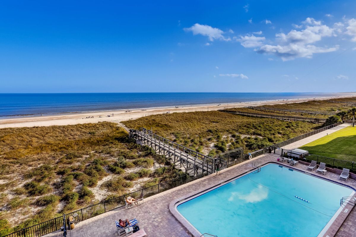 A swimming pool overlooks a sandy beach and ocean under a clear blue sky. A wooden boardwalk stretches over grassy dunes toward the shoreline. Lounge chairs surround the pool area.
