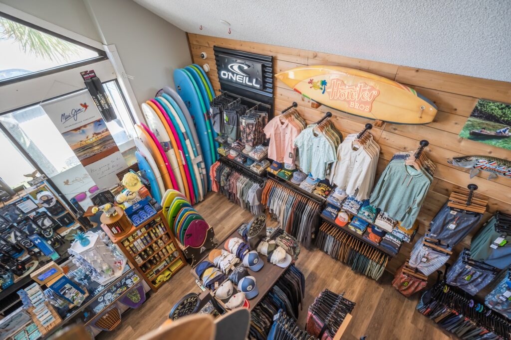 Aerial view of a surf shop displaying colorful surfboards, shirts, hats, and accessories neatly arranged on shelves and racks, with branded signs on the wooden walls.