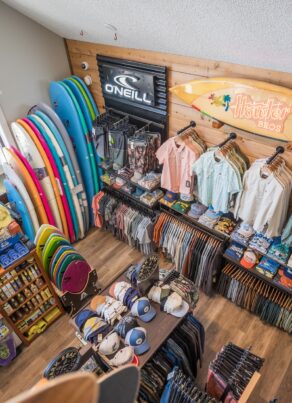 Aerial view of a surf shop displaying colorful surfboards, shirts, hats, and accessories neatly arranged on shelves and racks, with branded signs on the wooden walls.
