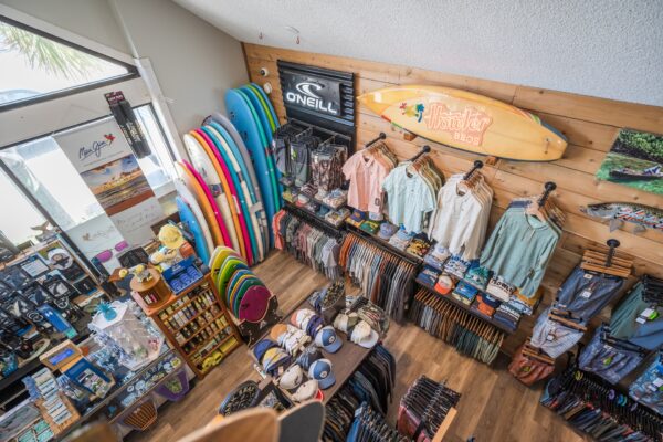 Aerial view of a surf shop displaying colorful surfboards, shirts, hats, and accessories neatly arranged on shelves and racks, with branded signs on the wooden walls.