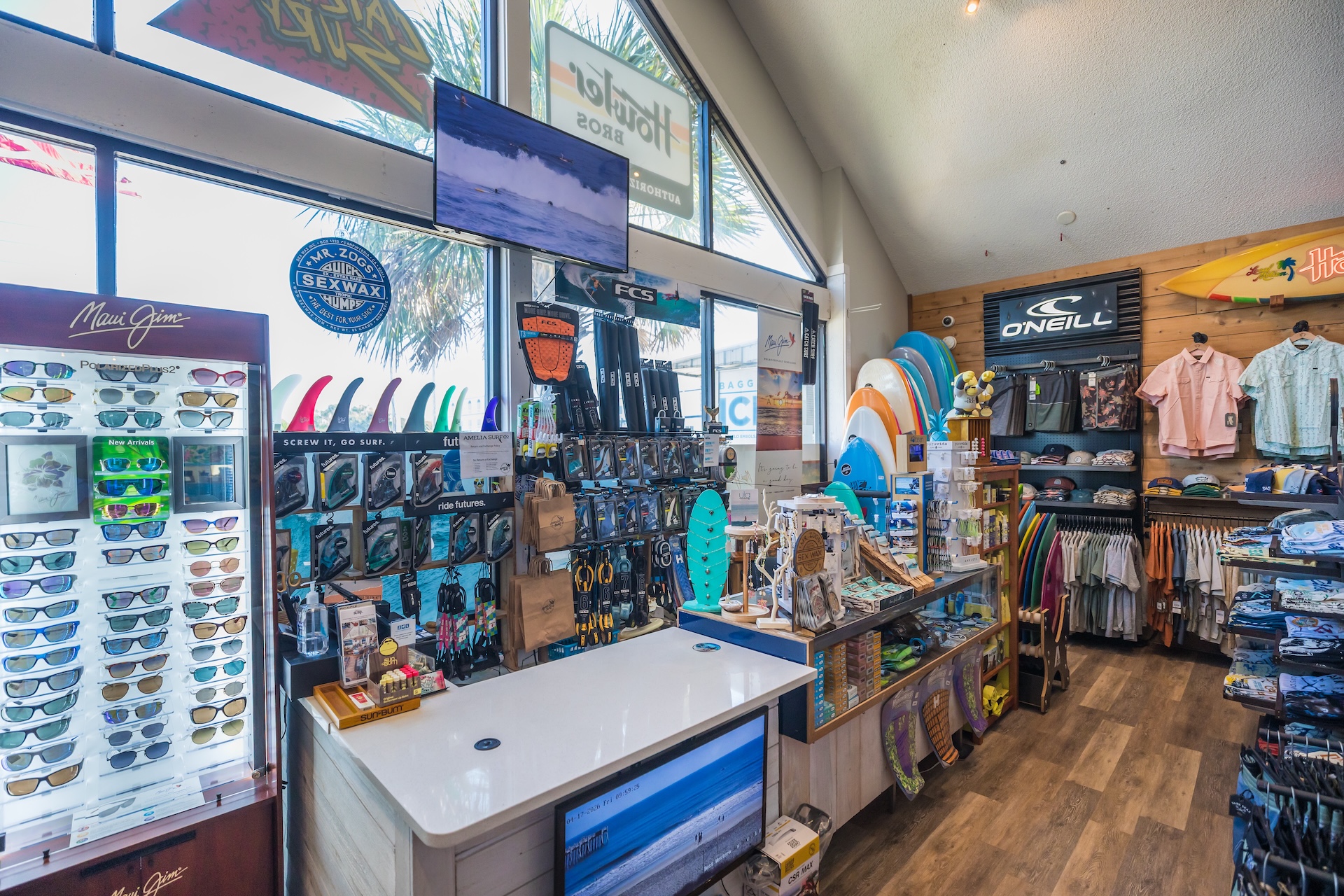 A surf shop interior with sunglasses, surfboards, wetsuits, hats, clothing, and accessories displayed on racks and shelves. Large windows let in natural light, and surfboards hang on the wall above the merchandise.