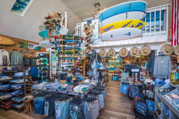 A surf shop interior with clothing racks, hats, accessories, skateboards, and several surfboards mounted on the upper wall. The store has a bright, beachy atmosphere with wooden floors and various merchandise neatly displayed.
