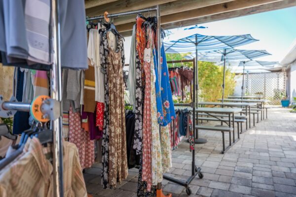 Clothing racks display dresses and garments under a shaded patio with blue umbrellas and picnic tables on a paved surface. The setting appears to be an outdoor market or pop-up shop.