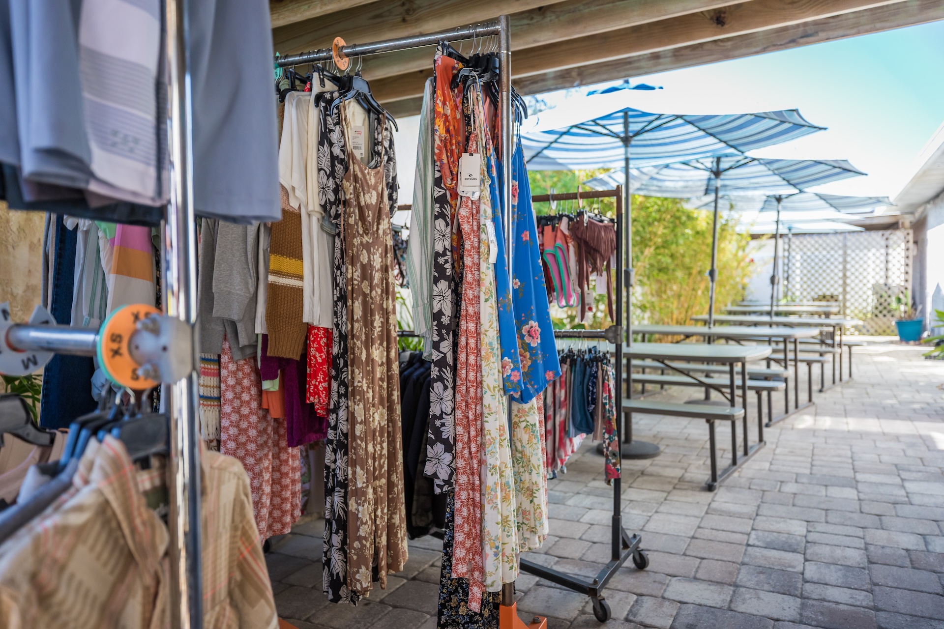 Clothing racks display dresses and garments under a shaded patio with blue umbrellas and picnic tables on a paved surface. The setting appears to be an outdoor market or pop-up shop.