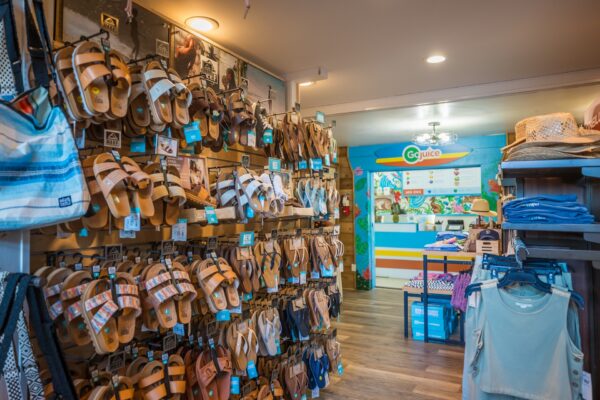 A retail store interior displays multiple pairs of sandals on a wall rack, with clothing, hats, and bags nearby. In the background, a colorful counter with a Go Juice sign is visible. The space has bright, inviting lighting.