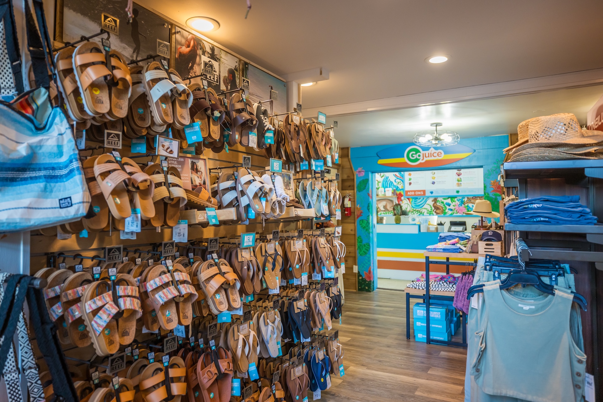 A retail store interior displays multiple pairs of sandals on a wall rack, with clothing, hats, and bags nearby. In the background, a colorful counter with a Go Juice sign is visible. The space has bright, inviting lighting.