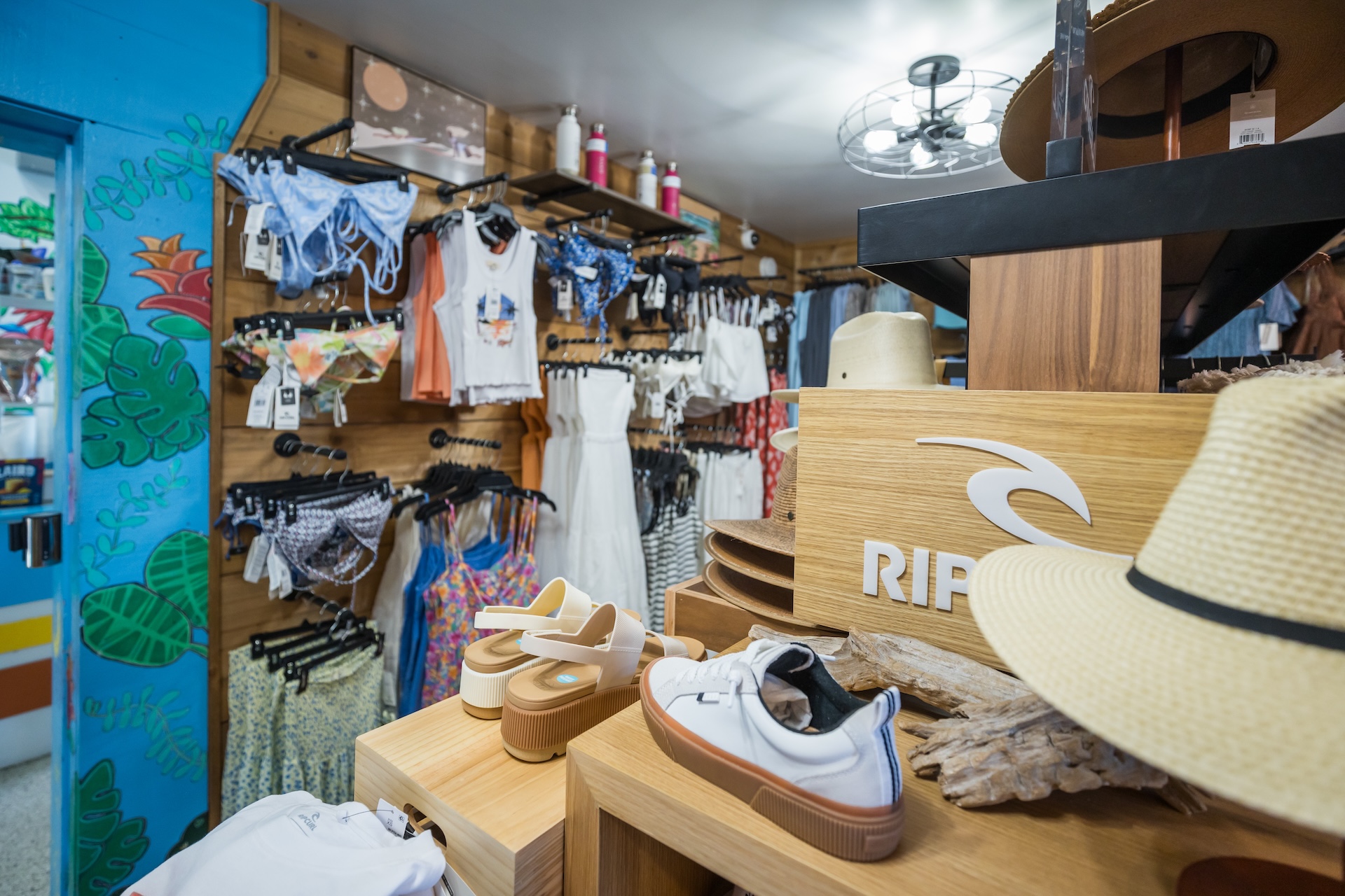 A shop interior displays swimwear, hats, sandals, and shirts neatly arranged on racks and shelves. A straw hat sits in the foreground next to Rip Curl shoes, with colorful clothing and beach accessories in the background.