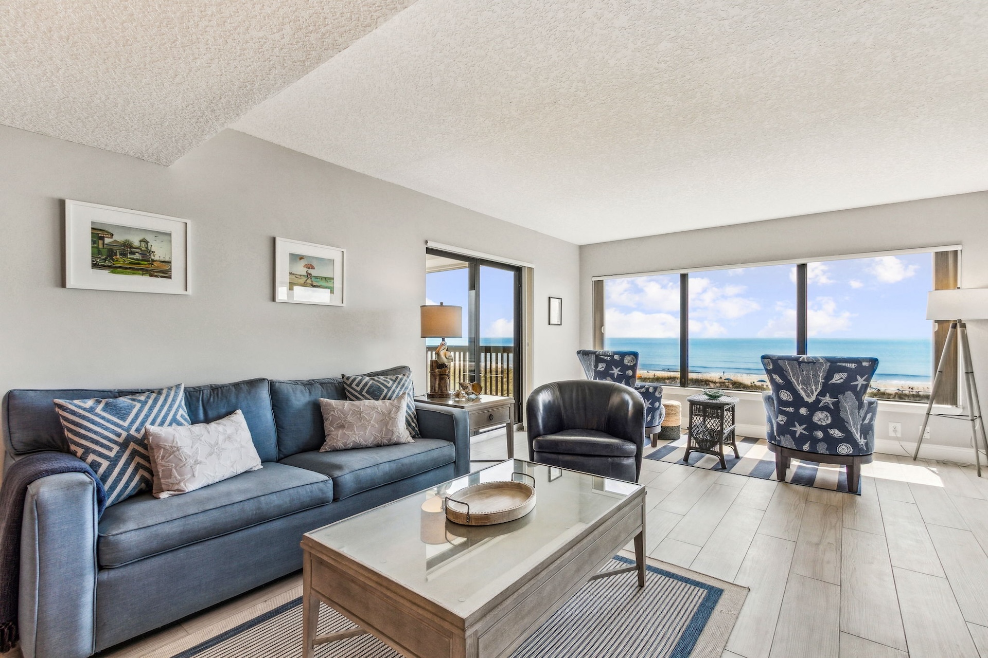 Bright, modern living room with a blue sofa, armchairs, coffee table, wall art, and large windows offering an ocean view under a sunny sky. Light wood floors and neutral décor create a coastal vibe.