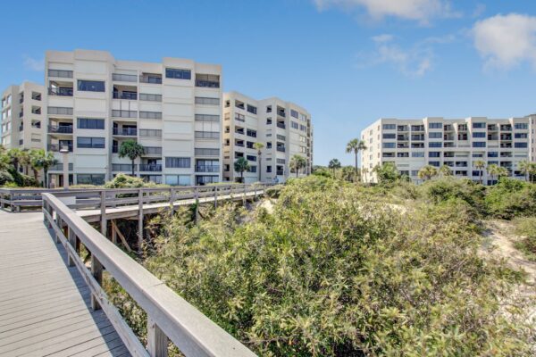 Two modern, white apartment buildings with balconies stand behind lush greenery, viewed from a wooden boardwalk under a blue sky with scattered clouds.