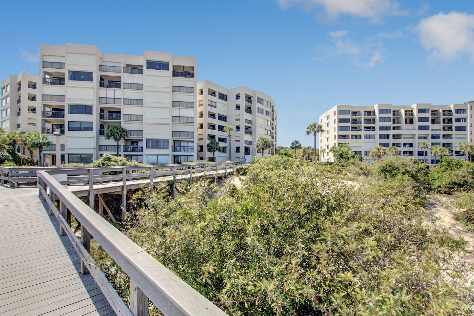 Two modern, white apartment buildings with balconies stand behind lush greenery, viewed from a wooden boardwalk under a blue sky with scattered clouds.