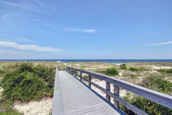 A wooden boardwalk leads through sand dunes and green shrubs toward a blue ocean under a clear sky.
