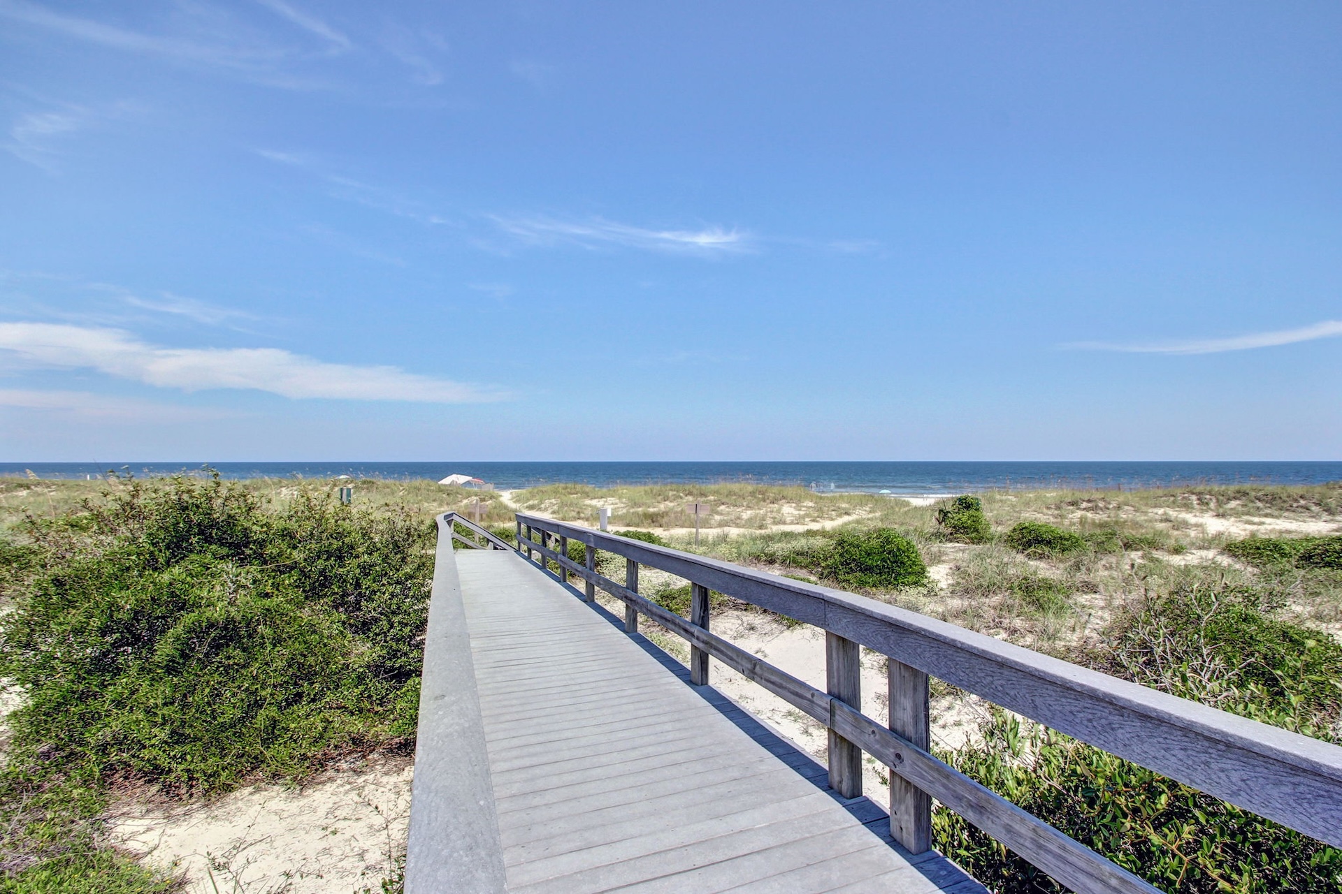 A wooden boardwalk leads through sand dunes and green shrubs toward a blue ocean under a clear sky.