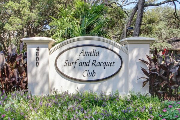 A sign reading Amelia Surf and Racquet Club is displayed on a white wall amid lush greenery, with purple flowers and dark-leaved plants in the foreground and the number 4800 on the left pillar.