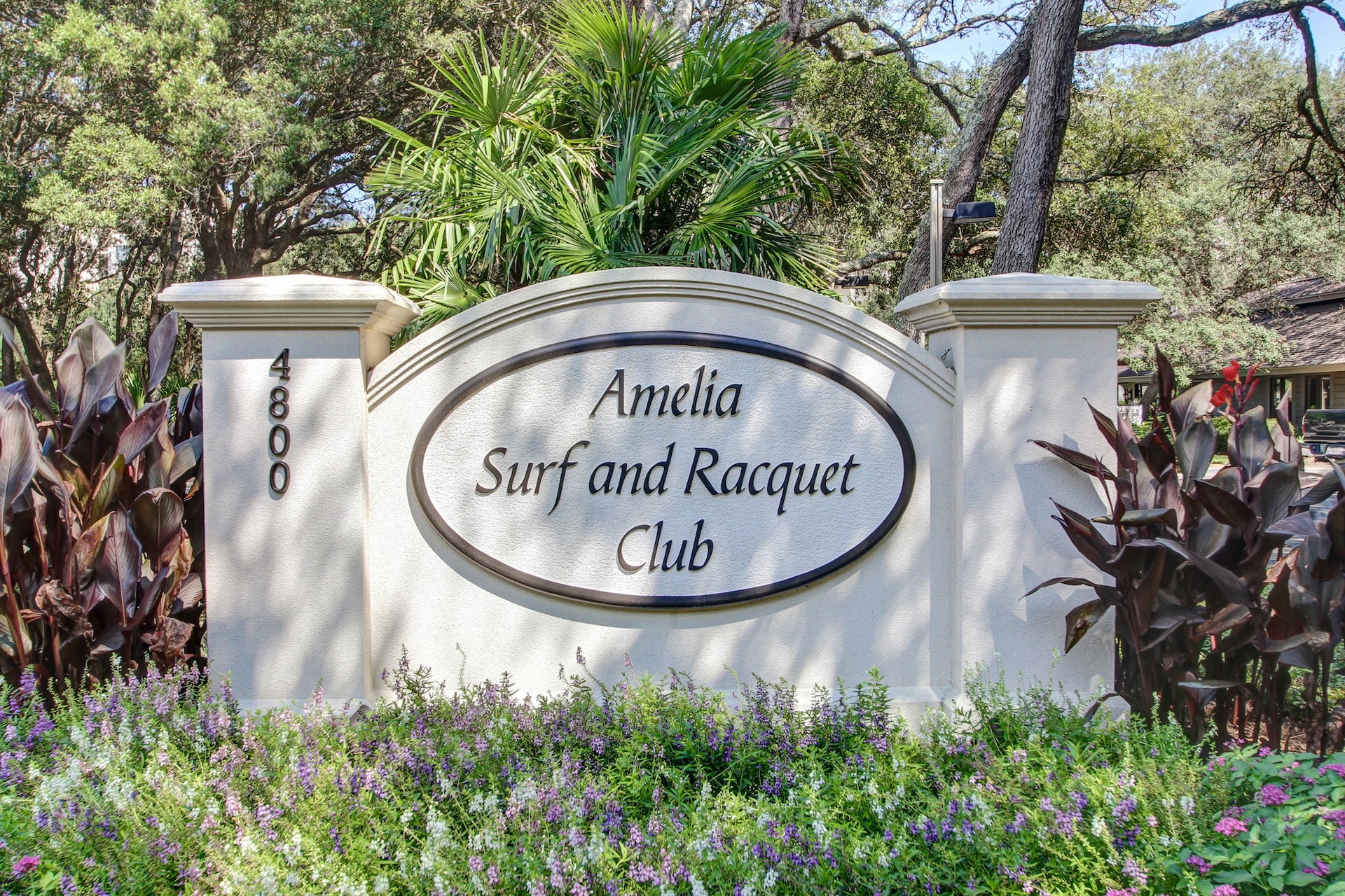 A sign reading Amelia Surf and Racquet Club is displayed on a white wall amid lush greenery, with purple flowers and dark-leaved plants in the foreground and the number 4800 on the left pillar.