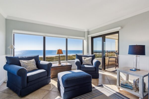 Bright living room with two blue armchairs, an ottoman, and large windows offering an ocean view. A side table with a blue lamp and a wooden chest are also in the room, with sunlight streaming in.