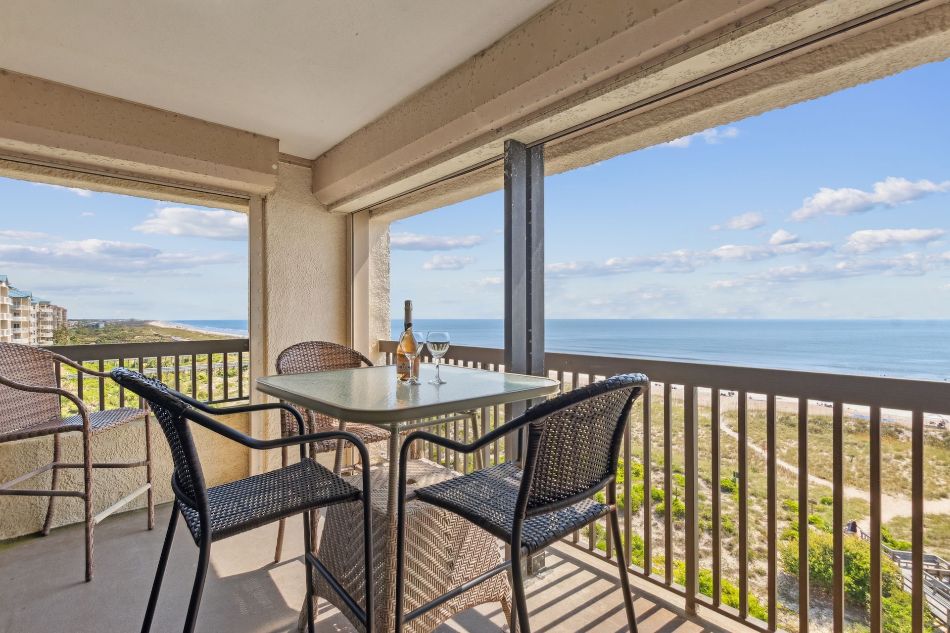 A balcony with a glass table, four wicker chairs, a bottle of wine, and glasses overlooks a beach, ocean, and blue sky with scattered clouds. Greenery and sand stretch below the balcony railing.