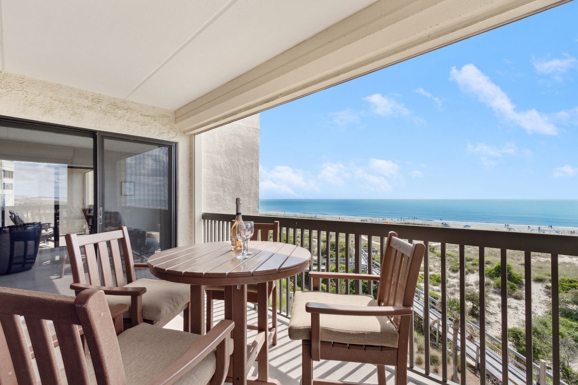 A balcony with a round wooden table, four chairs, a wine bottle, and two glasses overlooks a sandy beach and the ocean under a bright blue sky with scattered clouds.