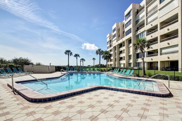 A sunny outdoor pool with turquoise lounge chairs, surrounded by a tiled deck. A modern multi-story building and palm trees are visible in the background under a clear blue sky.