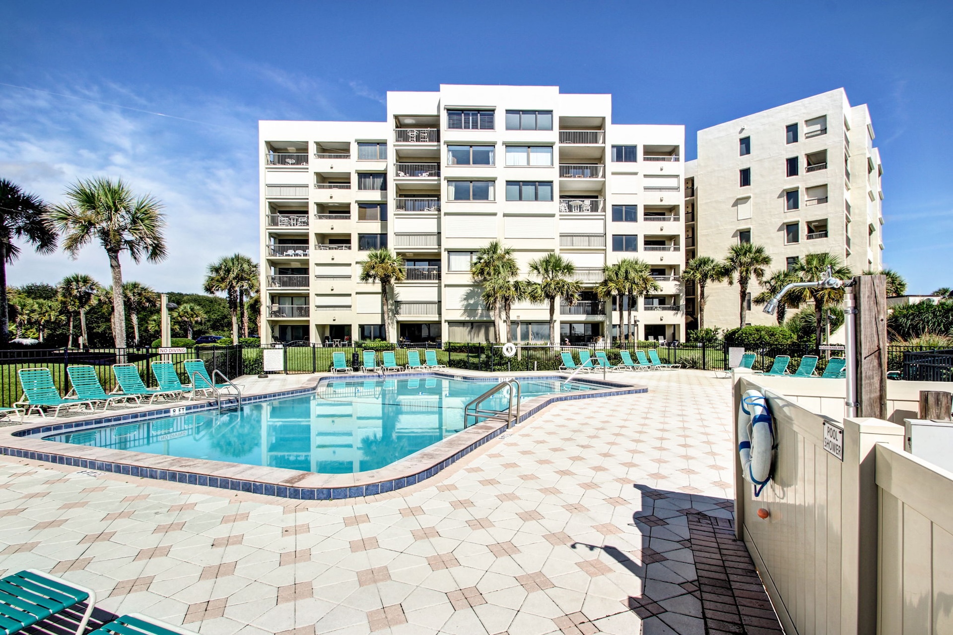 A clean outdoor swimming pool surrounded by green lounge chairs, palm trees, and a multi-story white apartment building under a clear blue sky.