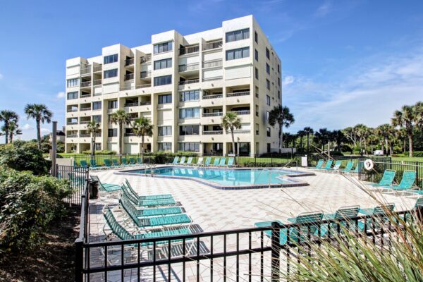 A modern white apartment building with balconies overlooks a fenced outdoor pool area, surrounded by teal lounge chairs and palm trees under a bright blue sky.