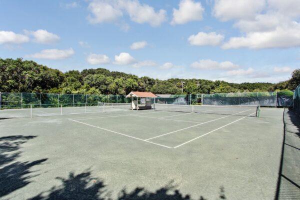 An empty outdoor tennis court with a clay surface, surrounded by green trees and a chain-link fence. A small shaded structure is near the center, and the sky is partly cloudy.