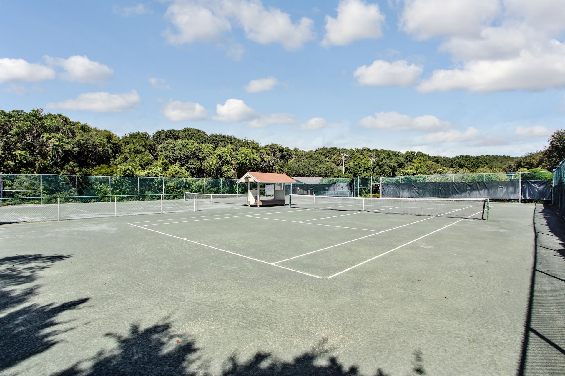 An empty outdoor tennis court with a clay surface, surrounded by green trees and a chain-link fence. A small shaded structure is near the center, and the sky is partly cloudy.