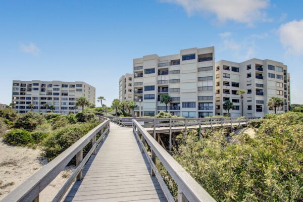 A wooden boardwalk leads through lush greenery toward two modern, multi-story beachfront condominium buildings under a bright blue sky with scattered clouds.