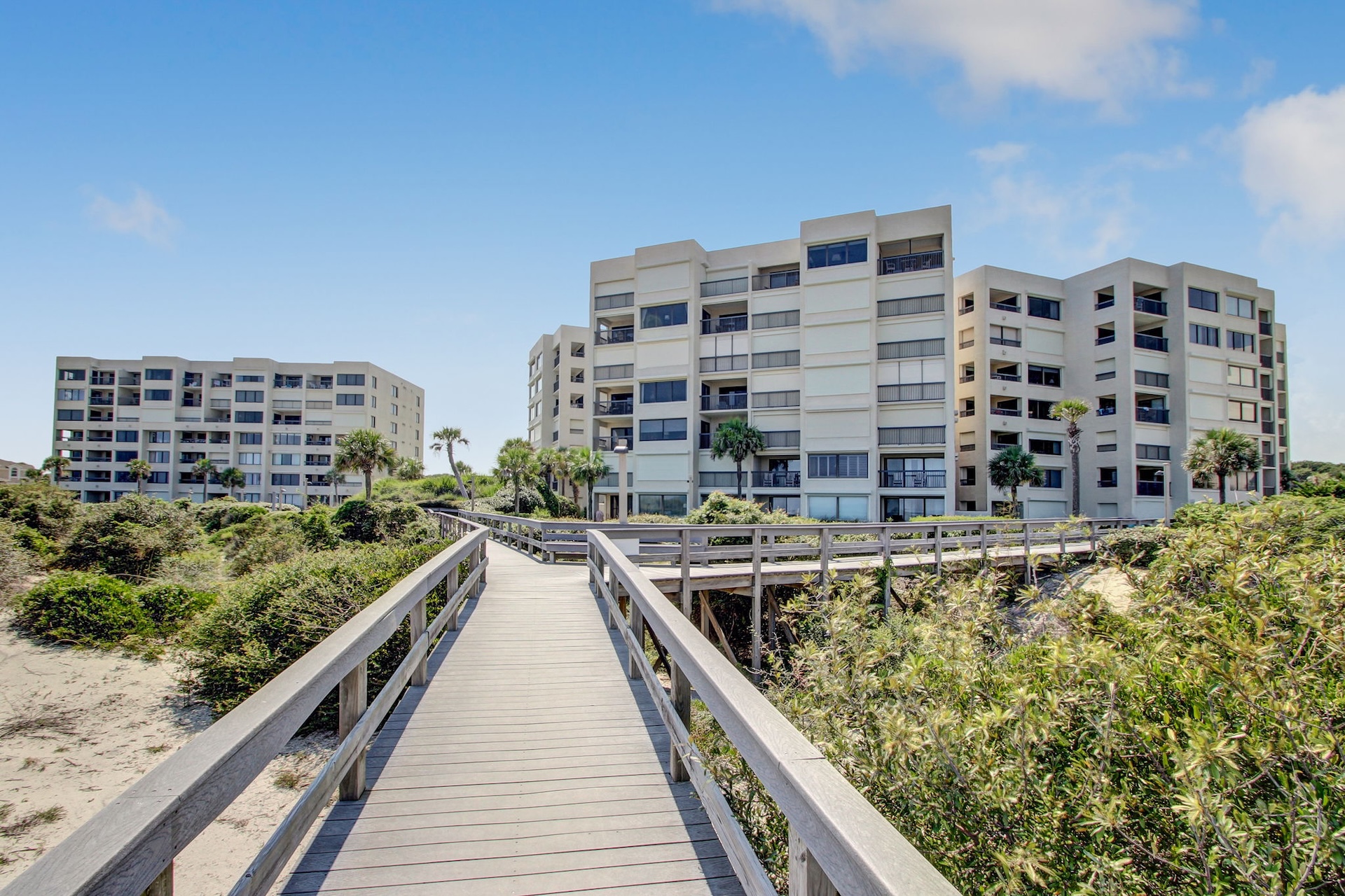 A wooden boardwalk leads through lush greenery toward two modern, multi-story beachfront condominium buildings under a bright blue sky with scattered clouds.