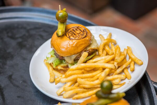 A cheeseburger topped with lettuce and cheese, served on a white plate with a generous portion of French fries. The burger bun is branded with ABC and is held together with a skewer and a green olive on top.