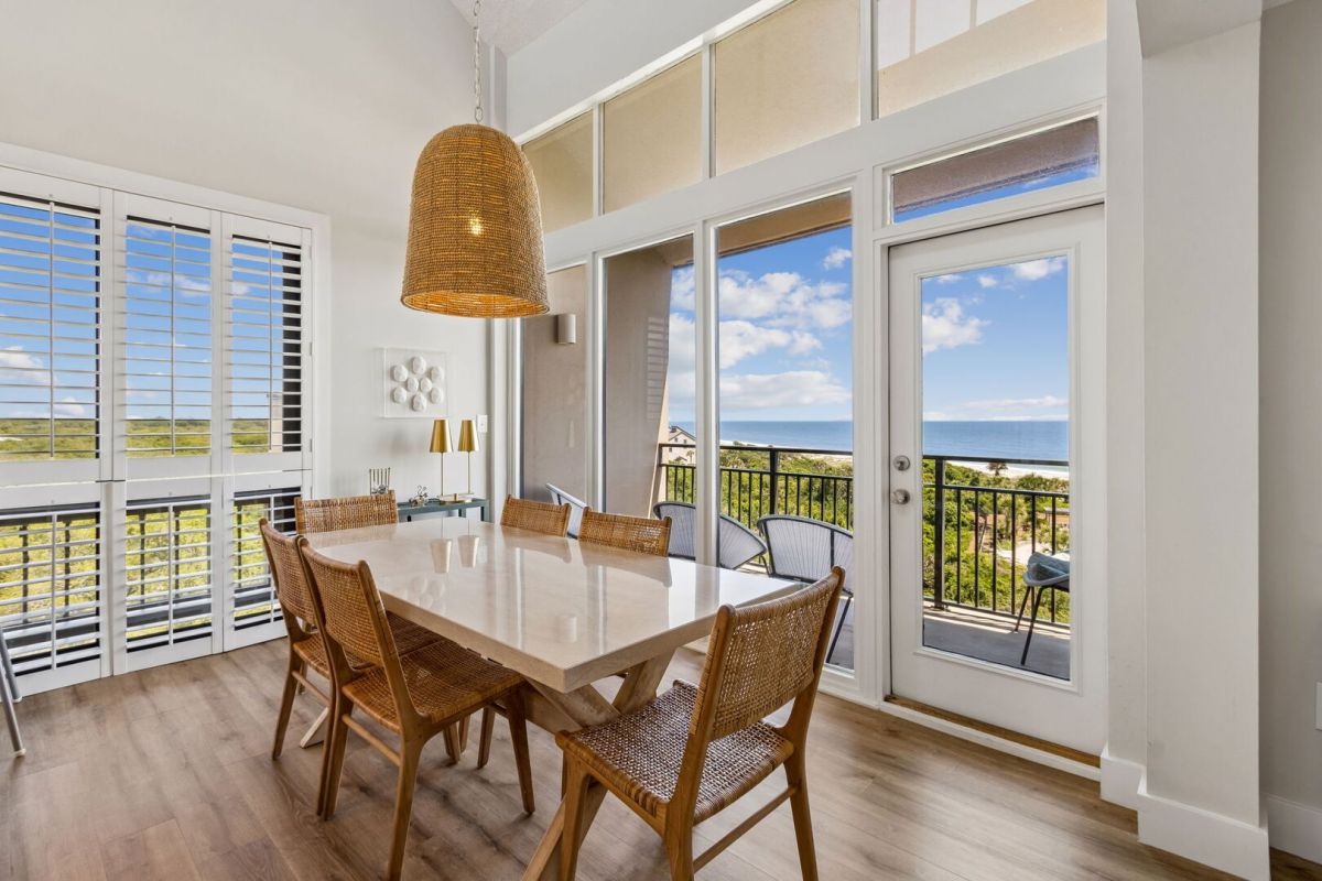 Dining area with six wicker chairs around a marble table, large pendant lamp above, wood flooring, and glass doors leading to a balcony with an ocean view under a blue sky with clouds.