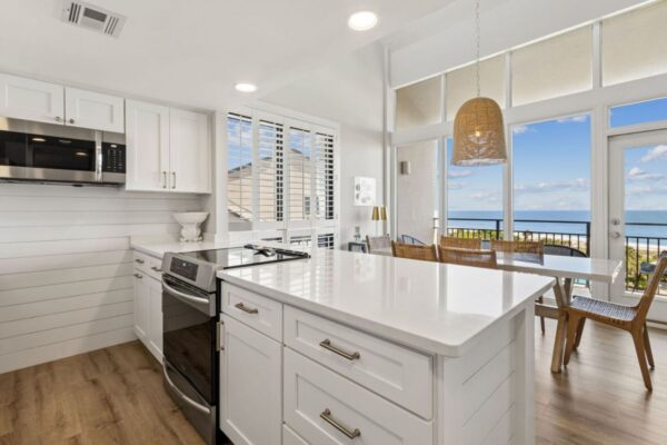 Bright kitchen with white cabinets, island, and stainless appliances. Dining area with wicker chairs and pendant light overlooks a balcony with an ocean view through large windows.