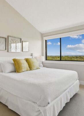 Bright bedroom with white bedding, two yellow pillows, white nightstands, lamps, framed art above the bed, and a large window showing a landscape with trees and blue sky with clouds.