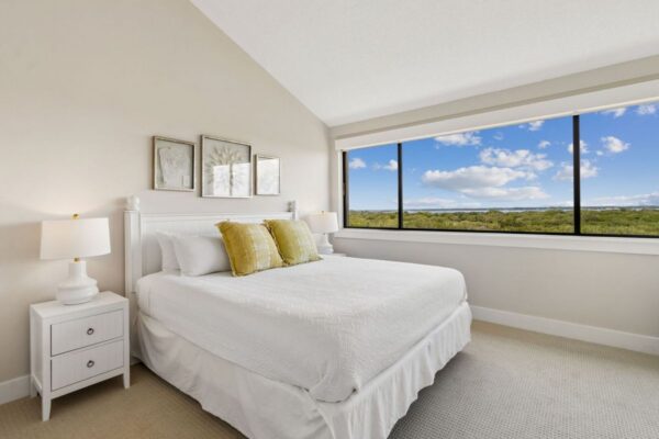 Bright bedroom with white bedding, two yellow pillows, white nightstands, lamps, framed art above the bed, and a large window showing a landscape with trees and blue sky with clouds.