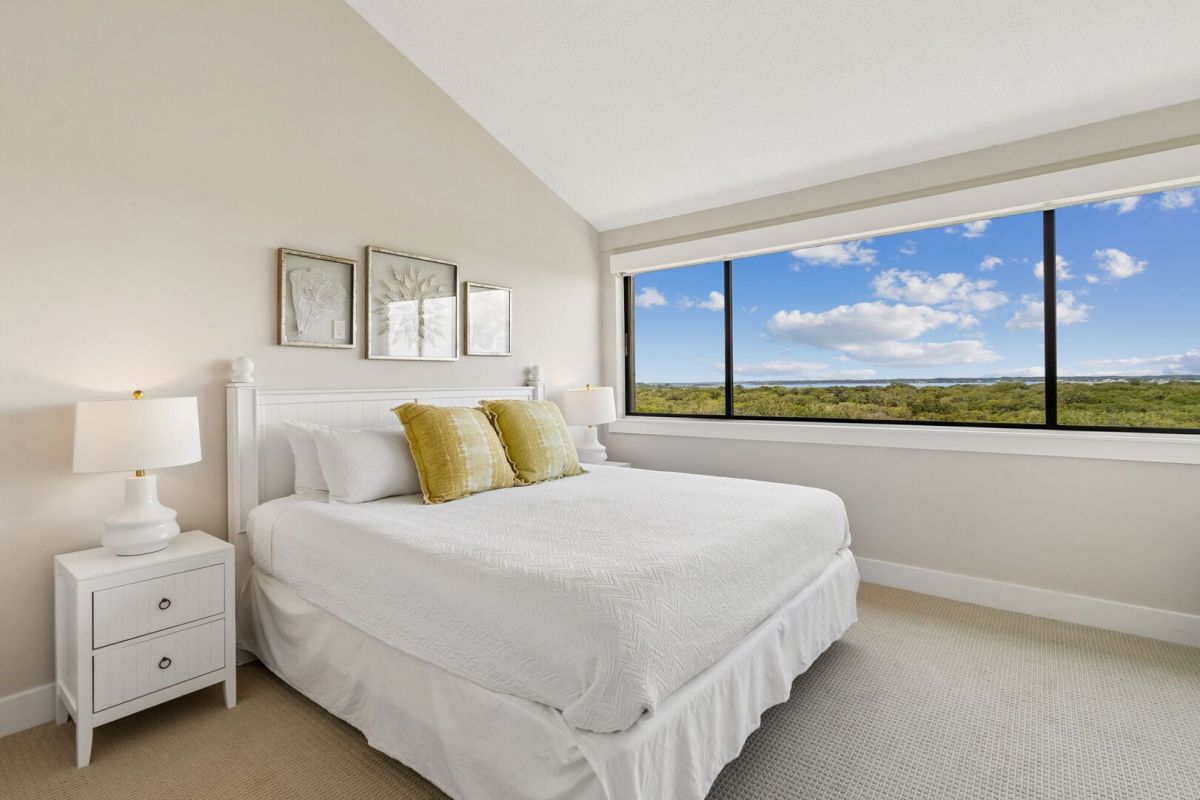 Bright bedroom with white bedding, two yellow pillows, white nightstands, lamps, framed art above the bed, and a large window showing a landscape with trees and blue sky with clouds.