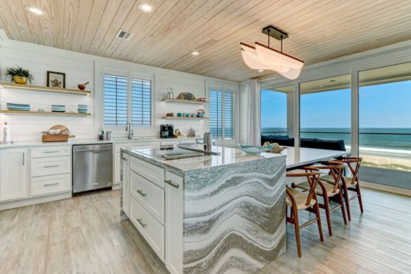 Modern beachside kitchen with a marble island, white cabinets, open shelves, wood ceiling, and large windows offering an ocean view. Four wooden chairs line the island, and natural light fills the space.