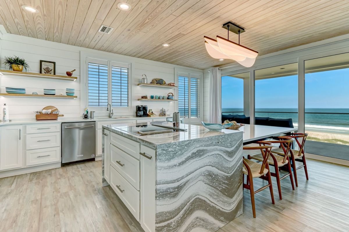 Modern beachside kitchen with a marble island, white cabinets, open shelves, wood ceiling, and large windows offering an ocean view. Four wooden chairs line the island, and natural light fills the space.