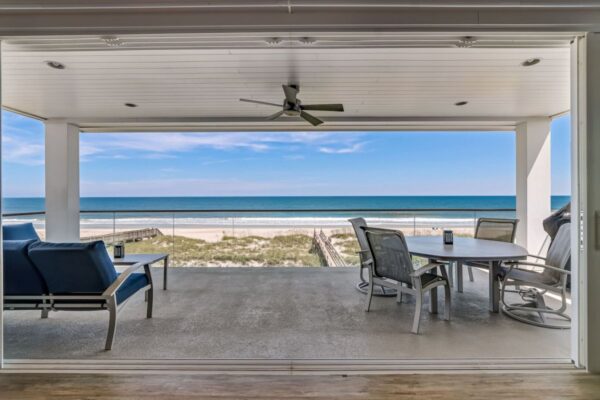 Covered patio with blue lounge chairs and a dining table overlooks sandy dunes and a calm ocean under a blue sky, seen through large open sliding doors.