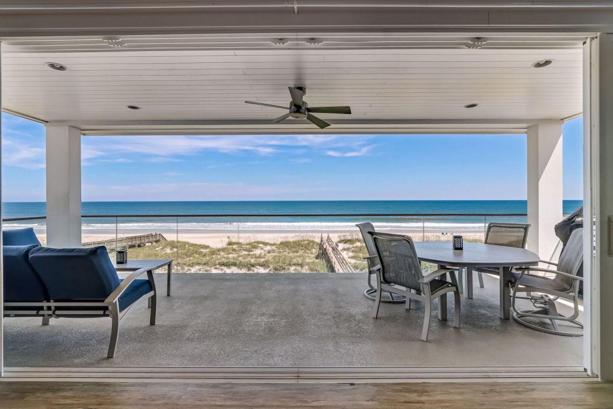 Covered patio with blue lounge chairs and a dining table overlooks sandy dunes and a calm ocean under a blue sky, seen through large open sliding doors.