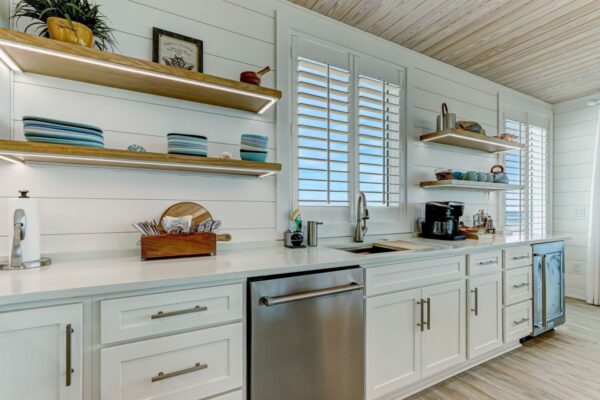 Modern kitchen with white cabinets, stainless steel dishwasher, sink, and open wooden shelves holding dishes and bowls. Window shutters let in natural light, and a coffee maker sits on the counter.