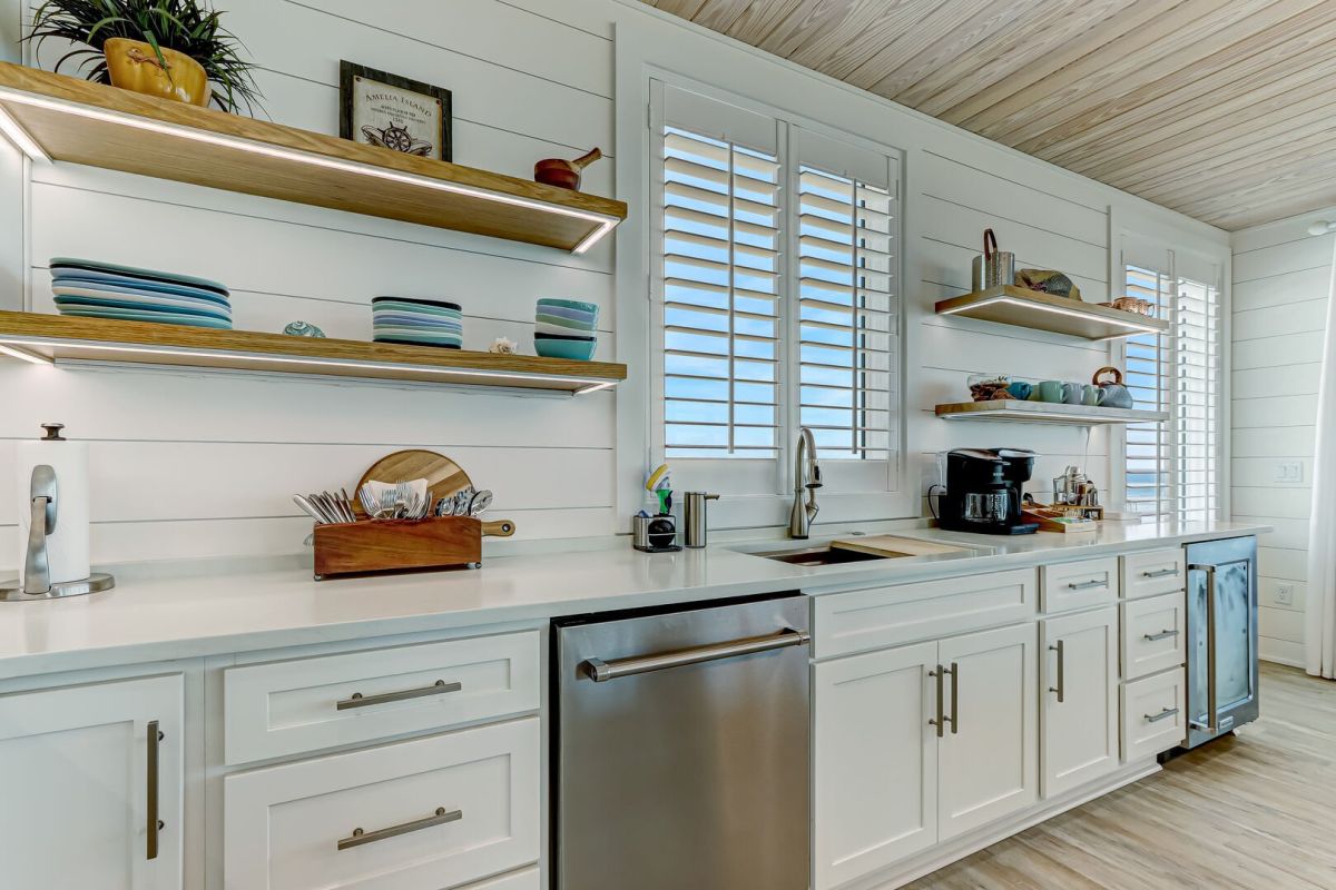 Modern kitchen with white cabinets, stainless steel dishwasher, sink, and open wooden shelves holding dishes and bowls. Window shutters let in natural light, and a coffee maker sits on the counter.