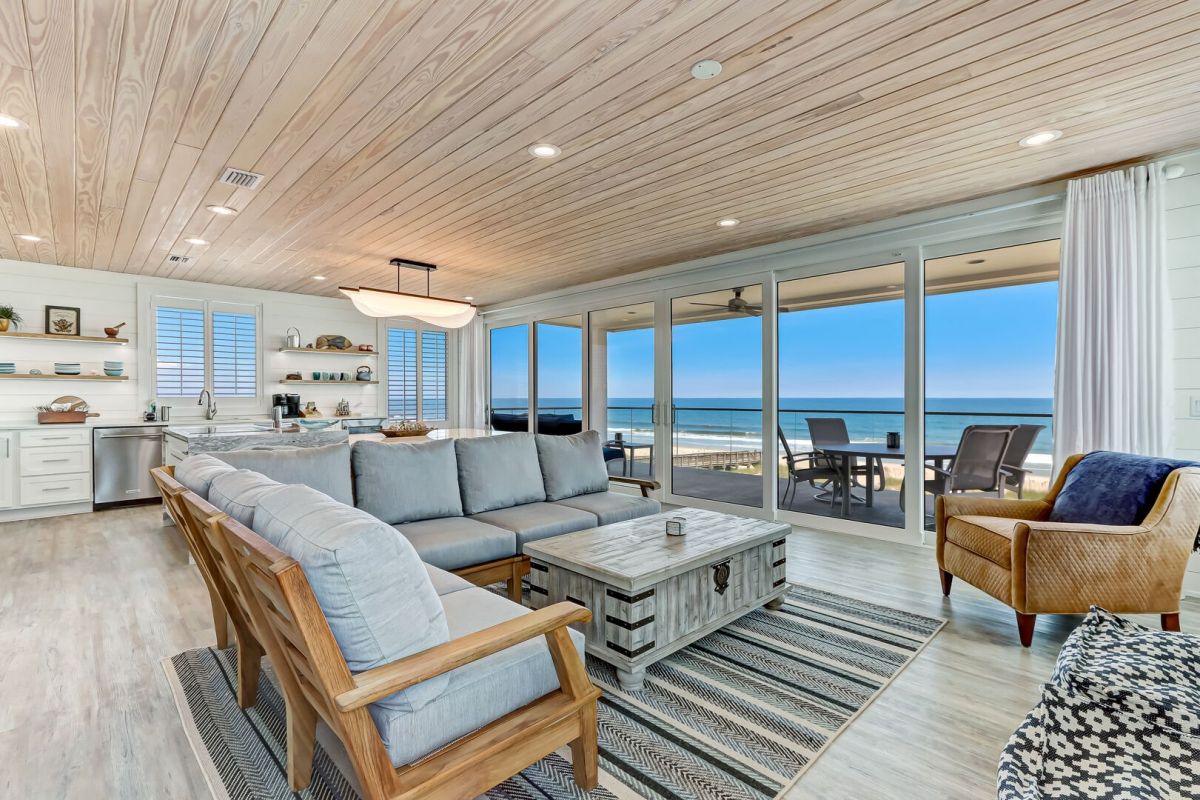 Modern beachfront living room with gray sofa, armchair, coffee table, and open kitchen. Floor-to-ceiling windows show an ocean view and lead to a patio with outdoor dining furniture.
