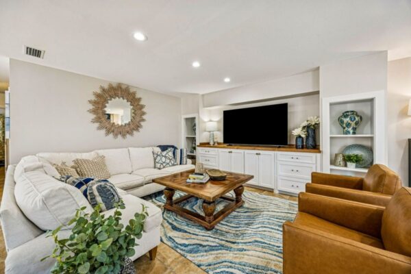 Bright living room with a white sectional sofa, two brown chairs, a wooden coffee table, TV on a white console, blue patterned rug, sunburst mirror, and decorative plants and vases.