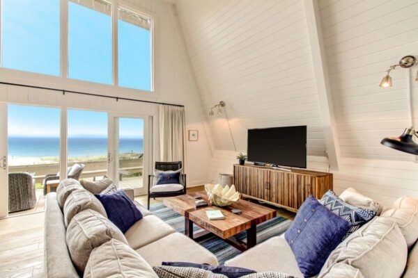 Bright living room with large windows showing an ocean view, a beige sectional sofa with blue pillows, a wooden coffee table, a TV on a console, and a striped rug on a light wood floor.