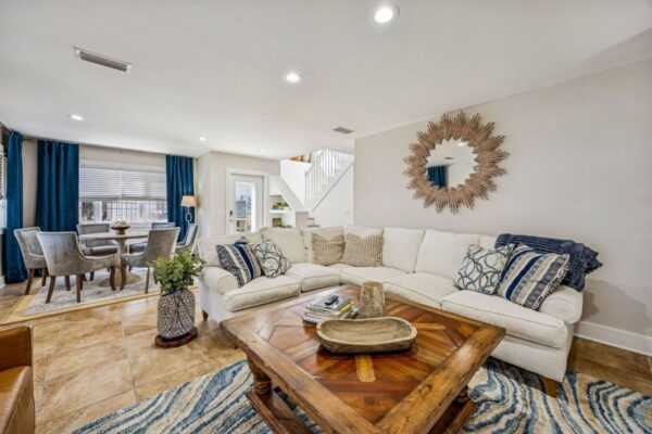Bright living room with a cream sectional sofa, wooden coffee table, patterned rug, and sunburst mirror. In the background, a dining area with a round table and blue curtains beside a staircase.