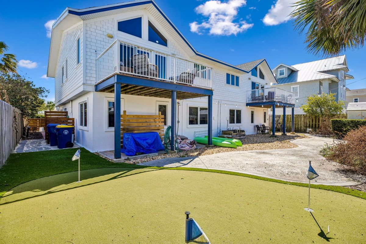Two-story white house with balconies, a covered patio, kayaks, and a small backyard putting green under a blue sky with scattered clouds. Palm trees and a neighboring house are visible.