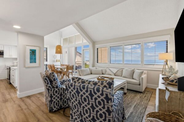 Bright coastal living room with patterned armchairs, white sectional sofa, large windows with ocean view, wooden dining table, and open kitchen. Light wood floors and beach-themed decor create a relaxing, airy atmosphere.